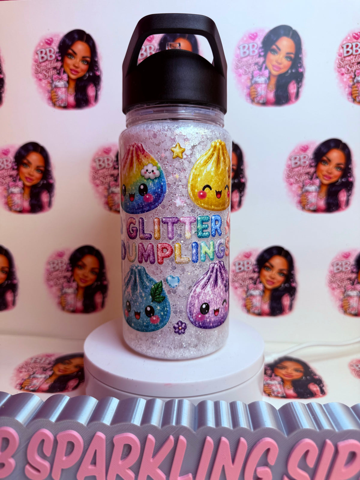 A glittery clear water bottle with a black lid, decorated with colorful ketchup and mustard illustrations and the words 'GLITTER DUMPLING'. The bottle is displayed on a white pedestal against a patterned backdrop featuring a repeating image of a woman.