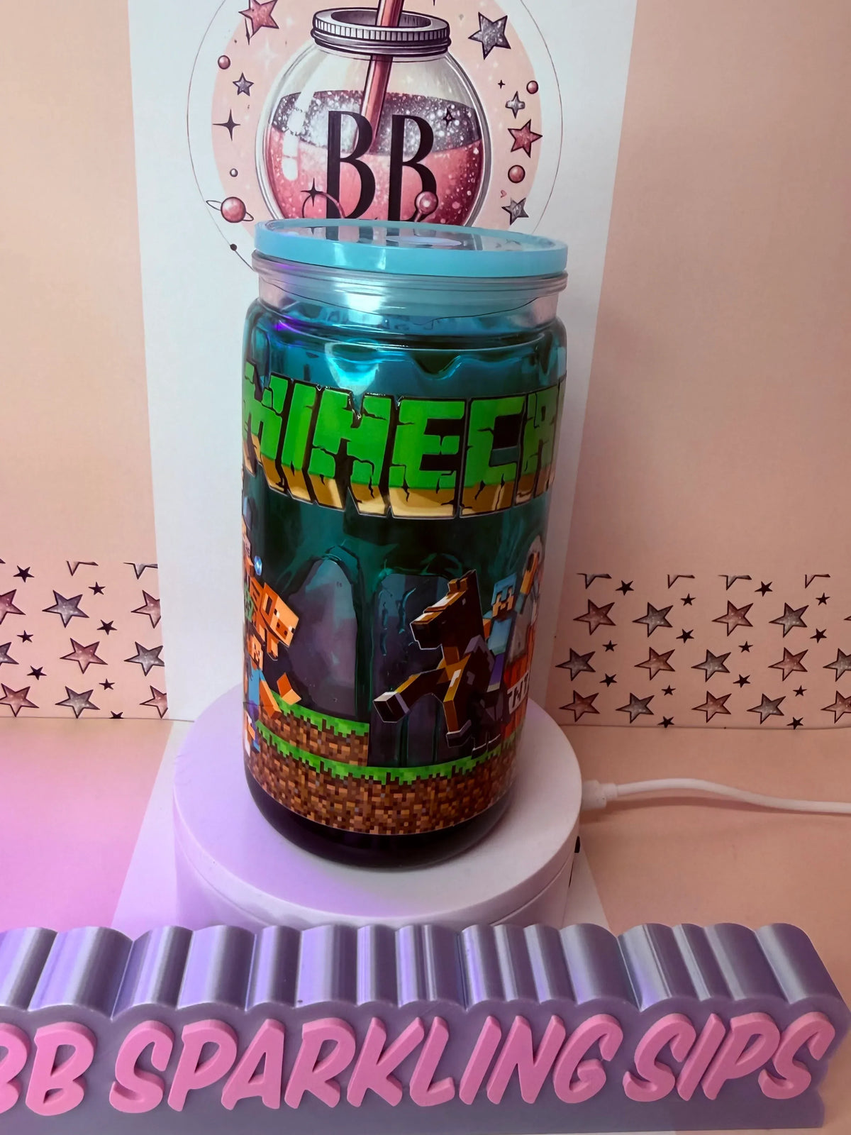 Minecraft-themed glitter tumbler with pixel art design and blue lid, displayed on a stand.
