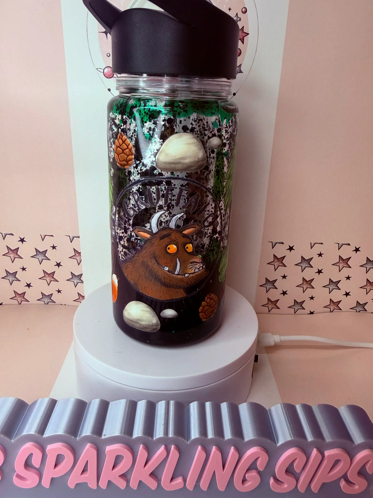 Handmade Gruffalo-themed glitter tumbler with pinecones, stones, and black lid, displayed on a stand.