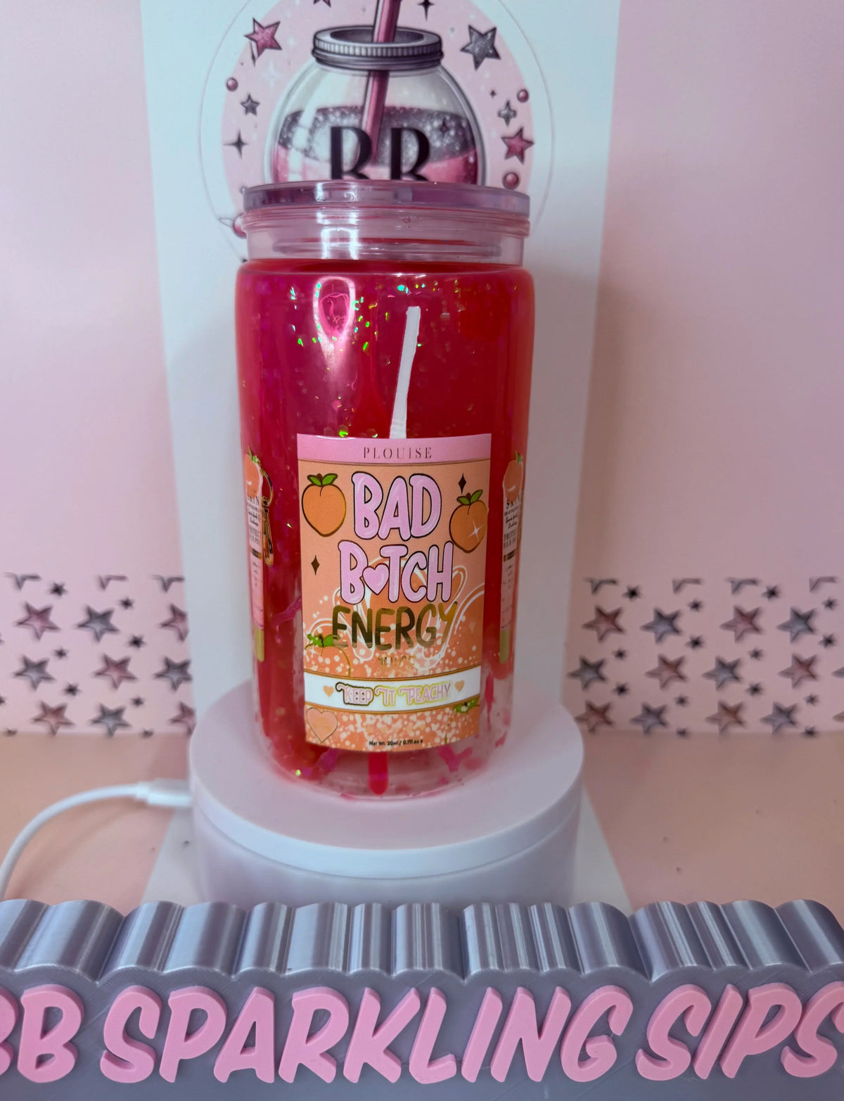 Pink glitter tumbler with 'Bad B*tch Energy' label, peach design, and straw on display stand